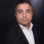The picture shows a man with short dark hair wearing a white shirt under a black blazer. He has a calm expression on his face and is standing in front of a dark, plain background.